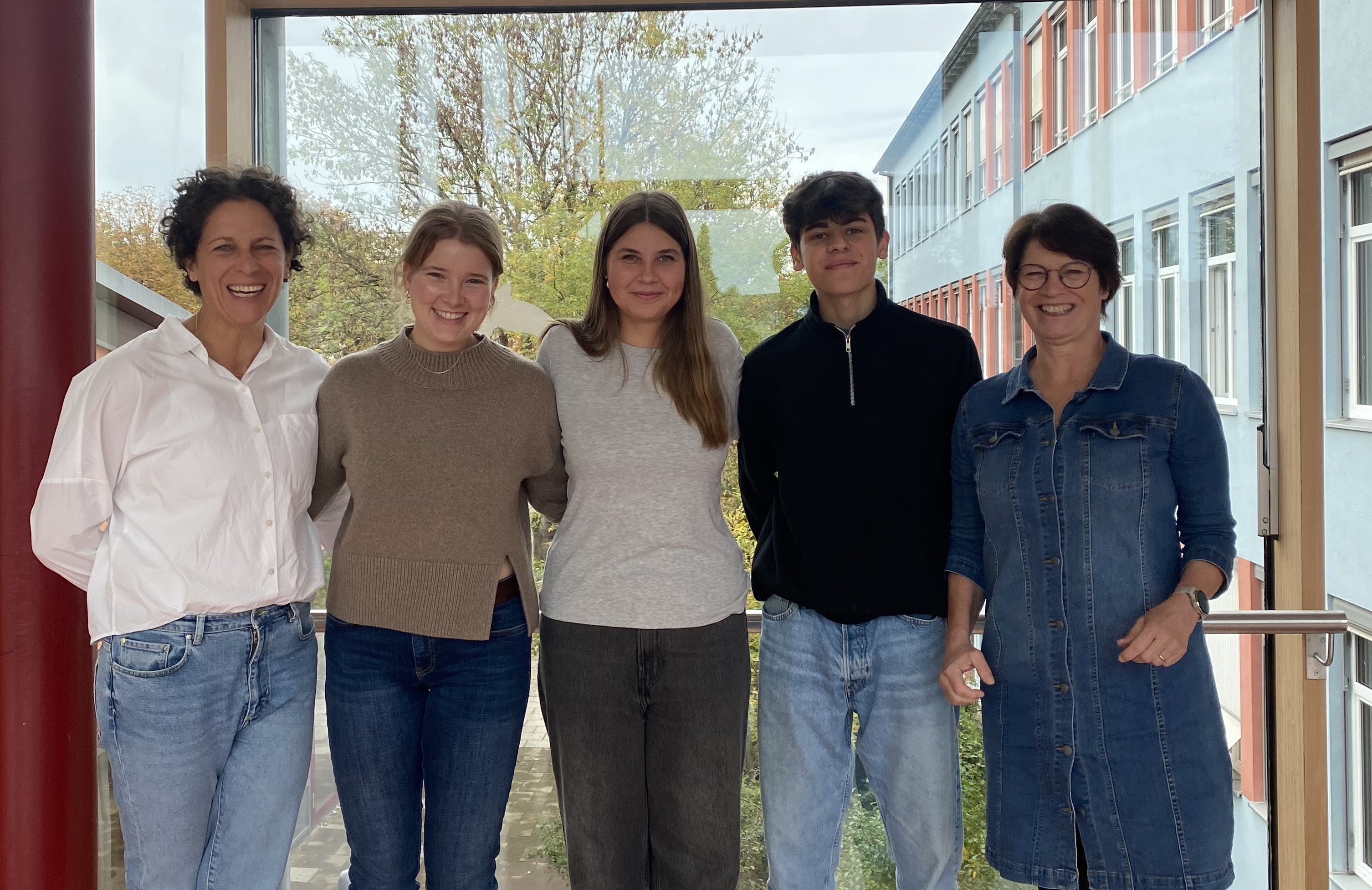 Die Betreuerinnen der SMV stehen rechts und links neben den zwei Schülersprecherinnen und dem Schülersprecher. Im Hintergrund ist durch ein Fenster ein Teil des Schulgebäudes sichtbar.