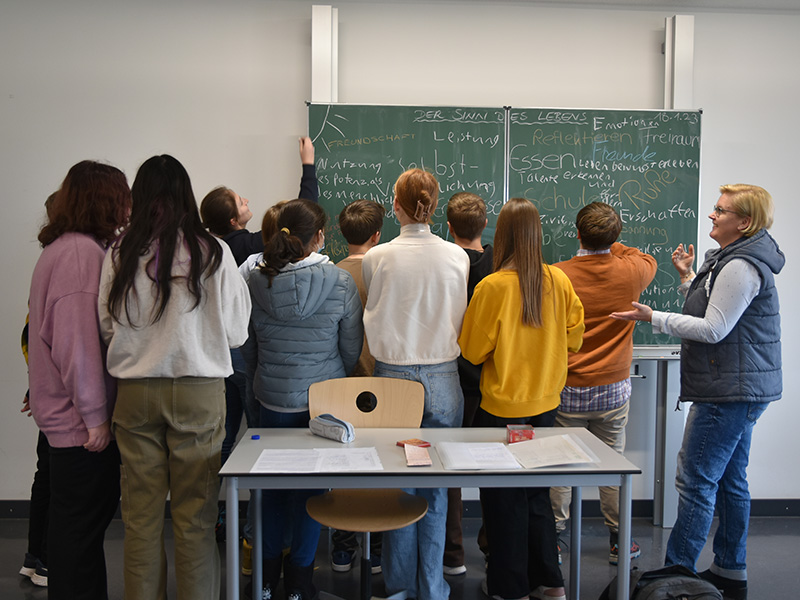 Schülerinnen stehen mit ihrer Lehrkraft vor einer Tafel. Sie schreiben Antworten zum Thema "Der Sinn des Lebens" auf die Tafel auf die Tafel..