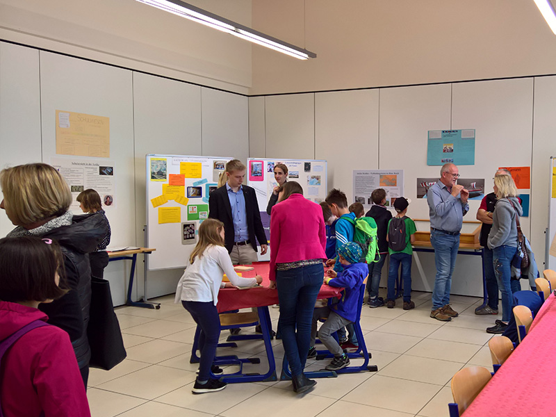 Schülerinnen und Schüler und Eltern betrachten Plakate zu Themen des Lateinunterrichts und machen Lateinspiele. an einem Tisch in der Mitte.