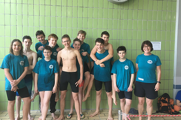 Schülerinnen und Schüler stehen mit ihrer Lehrkraft in einheitlicher Kleidung vor einer Schwimmbadwand. und strahlen in die Kamera.