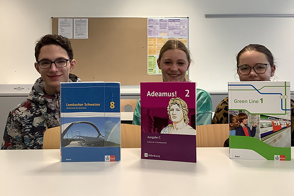 Zwei Schülerinnen und ein Schüler sitzen vor Schulbüchern für Mathematik, Latein und Englisch und lächeln freundlich in die Kamera..