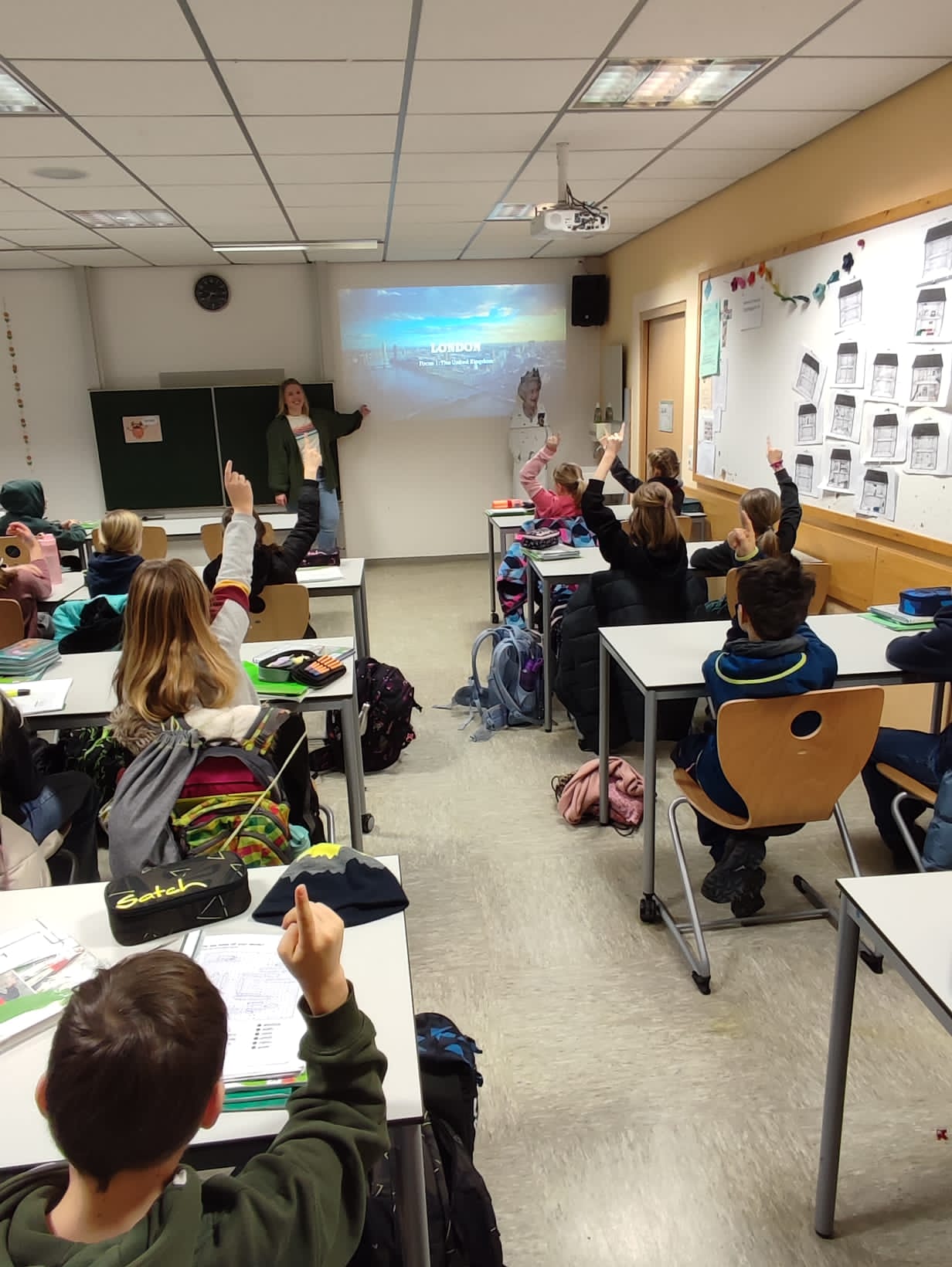 Eine Lehrerin erklärt vor einer Tafel und einem Londonbild. Schülerinnen und Schüler melden sich.