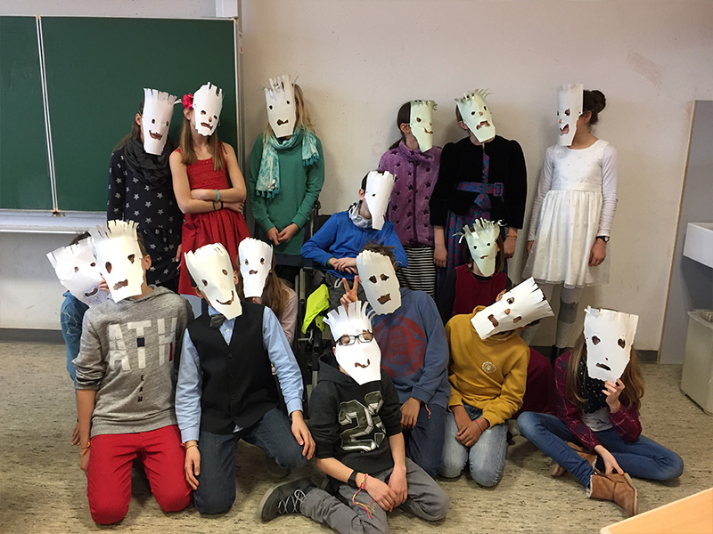 Eine Gruppe von Schülerinnen und Schülern posiert in einem Klassenzimmer. Alle haben Papiermasken auf.