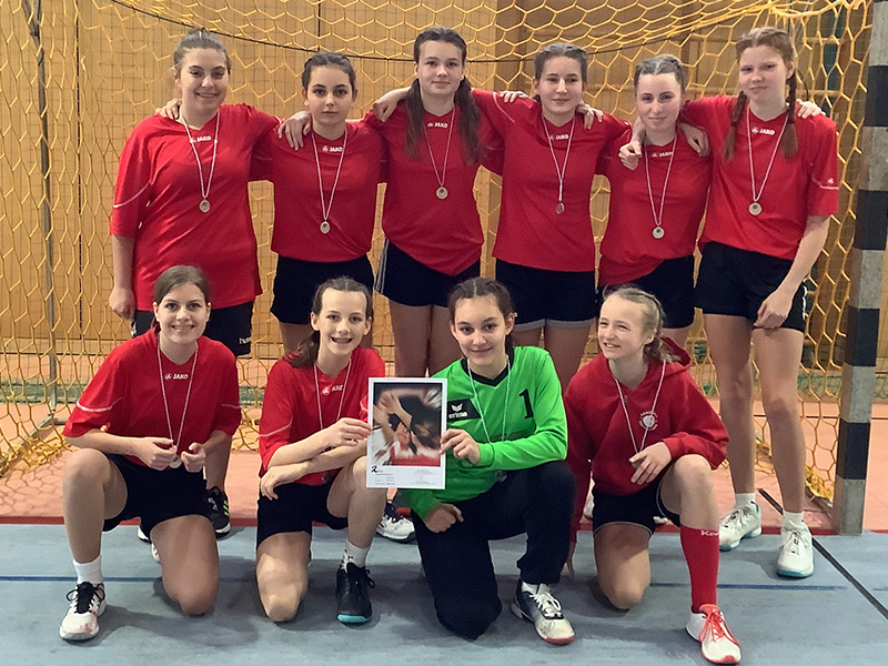 10 Handballerinnen tragen Medallien und posieren vor einem Handballtor für das Foto. Sie halten eine Urkunde.