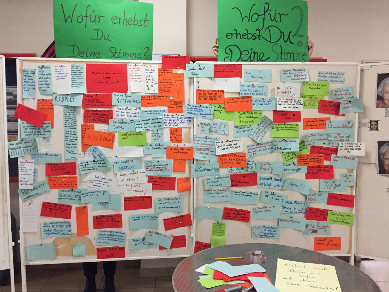 Zwei Stellwände sind zu sehen. Oberhalb der Stellwende sind grüne Poster mit der Überschrift "Wofür erhebst du Deine Stimme?" zu sehen. Auf den Stellwänden sind bunte Papiere mit Antworten auf die Frage angeheftet.
