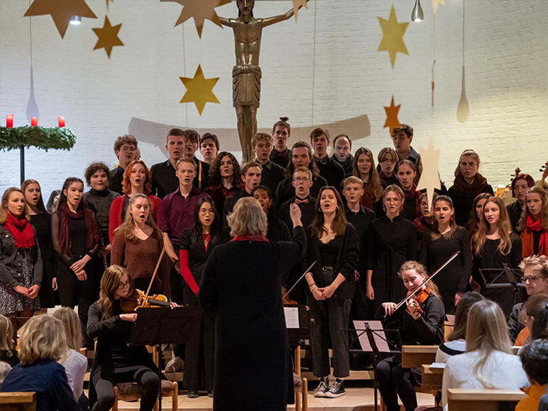 Schülerinnen und Schüler des Oberstufenchors singen in einer Kirche. Vor ihnen steht ihre Dirigentin, ebenso sitzt ein kleines Orchester vor ihnen.