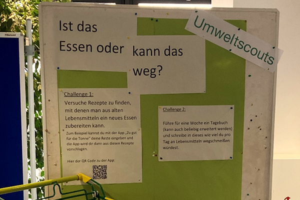 Vor einem grünen Hintergrund präsentiert die Posterwand provokante Fragen wie "Ist das Essen oder kann das weg?" sowie zwei Herausforderungen des Umweltscouts. Unten links im Bild sieht man einen Einkaufswagen.