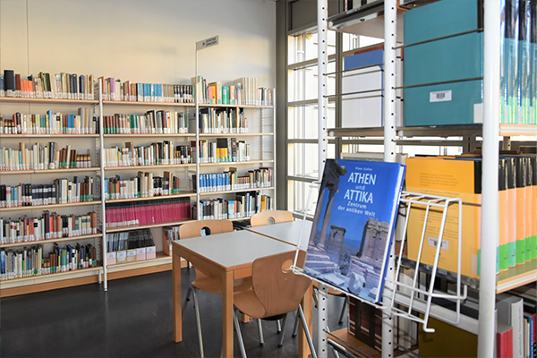 Im Hintergrund ist ein gut sortiertes Bücherregal sichtbar, während in der Mitte zwei Tische und Stühle zum Verweilen einladen. Im Vordergrund hebt sich das Buch „Athen und Attila“ hervor.