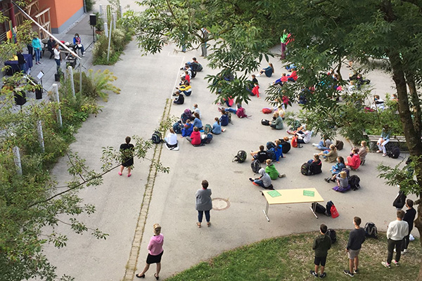 Von oben fotografiert, bietet sich ein Blick durch die Bäume auf den Pausenhof. Vorne versammeln sich Kinder, die dem Jahresabschlussgottesdienst aufmerksam lauschen. 