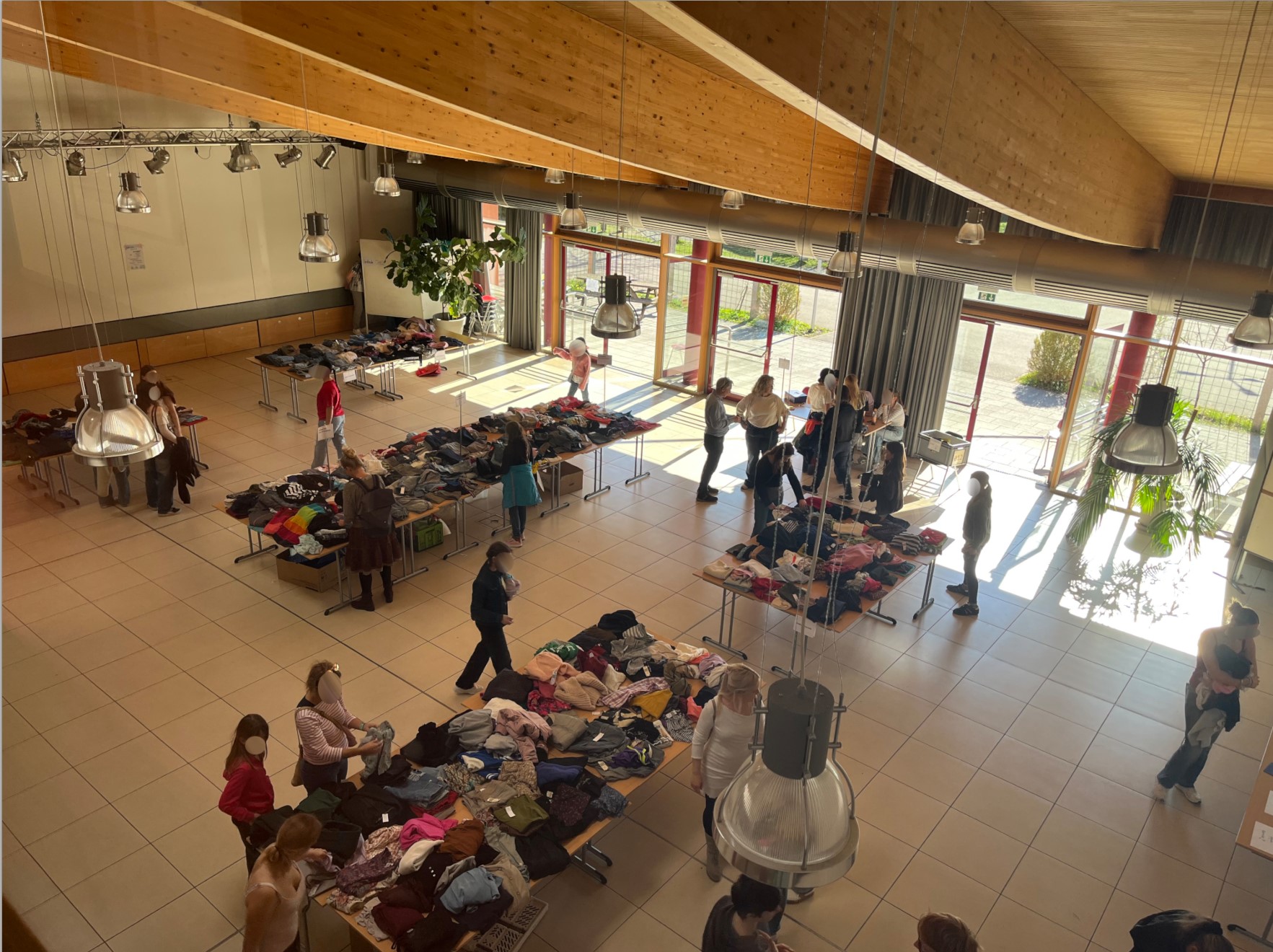 Zu sehen ist die Schulaula. Auf Schultischen, die zu Marktständen umgewandelt sind, ist Kleidung drapiert, durch welche die Besucherinnen und Besucher stöbern.