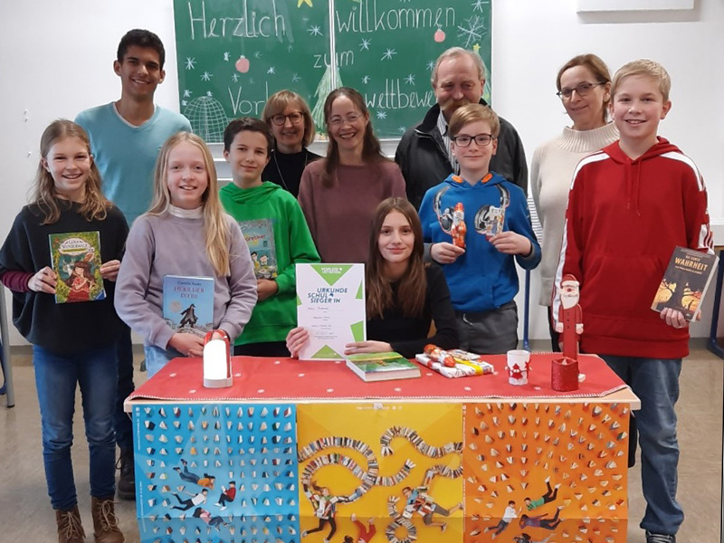Kinder halten ihre Preise des Lesewettbewerbs und strahlen - neben ihren Lehrkräften und ihrer Direktorin - in die Kamera.