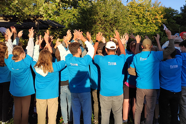 Jugendliche Schülerinnen und Schüler, gekleidet in blauen T-shirts mit der Aufschrift "Tutor" stehen mit dem Rücken zur Kamera und halten ihre Hände in die Höhe.