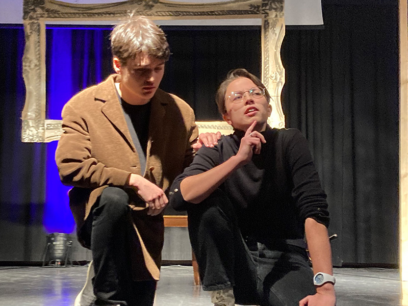 Zwei Schüler knien vor einem Bildrahmen auf einer Bühne und spielen Theater.